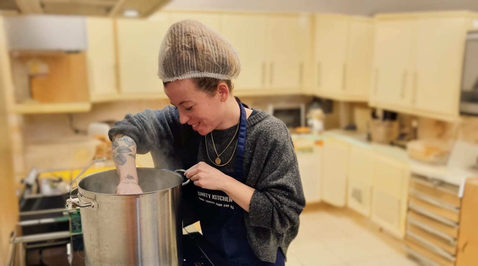 Harrie, one of the Community Kitchen Trustees, prepares a large pan of steaming food on the hob in a working kitchen.