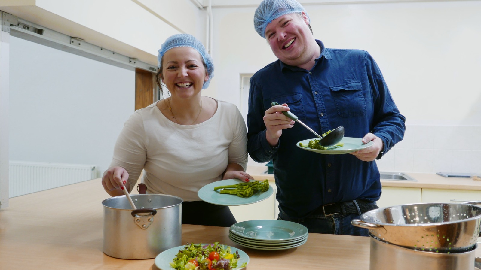 Picture of Molly and Jon, two of Community Kitchen's trustees serving food behind a kitchen serving hatch